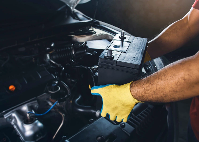 The image illustrates a technician carefully lifting a unit while performing a car battery change. The focus on the yellow gloves and firm grip highlights the proper handling and safety techniques required when working in the engine compartment.